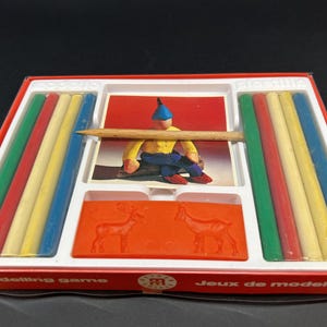 May include: A rectangular modelling game set in a red box containing eight cylindrical wax sticks in green, red, yellow, and blue. The set includes a central card depicting a figure, a wooden tool, and an orange rectangular mold with deer.