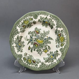 May include: A round ceramic plate with a cream-colored center and a wide green border. The design features a central floral arrangement with yellow and pink flowers, green foliage, and two small birds. The border has a textured green pattern.