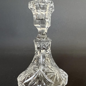 May include: A clear glass candlestick holder featuring a faceted, geometric stem and a wide, flared base. The base is decorated with a pressed starburst pattern, while the top section has a hexagonal opening designed to hold a single taper candle.