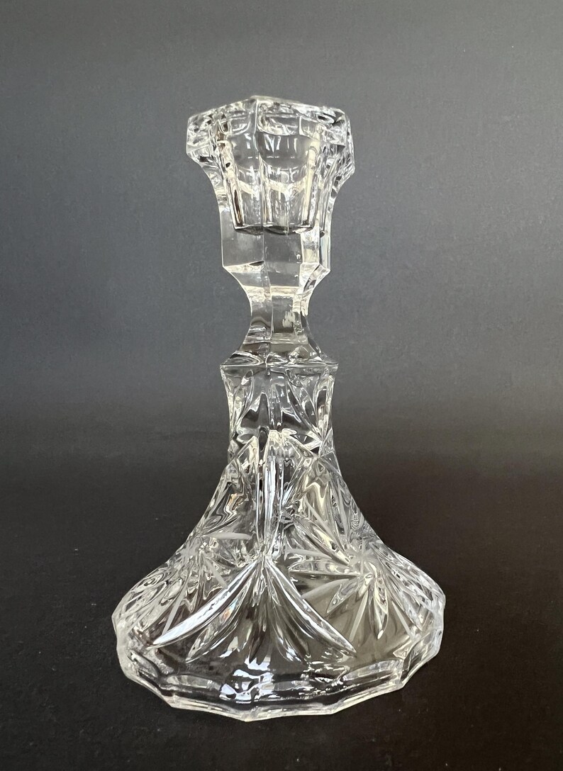 May include: A clear glass candlestick holder featuring a faceted, geometric stem and a wide, flared base. The base is decorated with a pressed starburst pattern, while the top section has a hexagonal opening designed to hold a single taper candle.