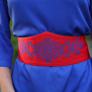 May include: A blue dress with a red belt. The belt features a blue embroidered floral design. The sleeves are three-quarter length. The background is blurred.