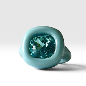 May include: A ring featuring a smooth, rounded, light blue band with a glossy finish. The center is set with a large, square-cut, faceted teal gemstone that reflects light, creating a contrast between the matte-like band and the crystal.