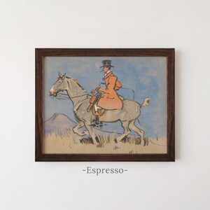 May include: A framed print featuring a stylized illustration of a rider in a top hat and orange coat mounted on a dappled gray horse. The scene includes a blue sky and a tan landscape. The word -Espresso- is printed below the dark wood frame.
