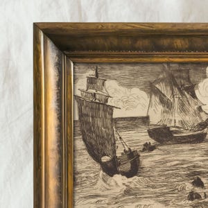 May include: A framed nautical etching depicts two sailing ships at sea. The artwork is in black and white, with detailed linework showing the waves and clouds. The frame is a warm gold color, adding a classic touch.