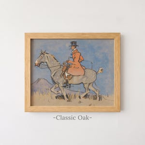 May include: A framed print in a light-colored wooden frame featuring an illustration of a person in an orange coat and top hat riding a grey horse. The scene includes a blue sky and a tan landscape. The text -Classic Oak- is printed below the frame.