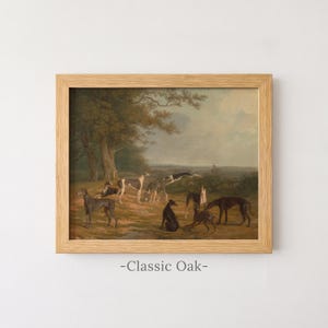 May include: A framed landscape painting featuring a group of greyhounds in a field with trees under a cloudy sky. The dogs are depicted in various poses, including standing and lying down. The artwork is enclosed in a light-coloured wooden frame with the text -Classic Oak- printed below the frame.