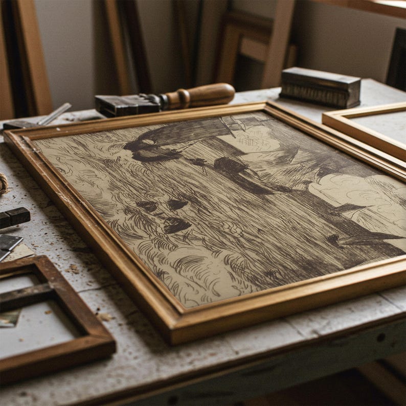 May include: A framed sepia-toned artwork with a gold-colored frame rests on a work table. The artwork depicts a detailed sketch of a person in a field. Framing tools and other framed pieces are visible in the background, suggesting a workshop setting.