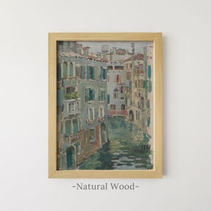 May include: A framed oil painting depicting a canal scene in Venice with multi-story buildings in shades of grey, blue, and muted red, reflected in the water. The artwork is dated 1888 and is encased in a light-colored natural wood frame.