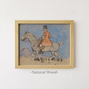 May include: A framed print in a natural wood frame featuring an illustration of a rider in a top hat and orange coat mounted on a grey dappled horse. The scene is set against a blue sky and a landscape with a mountain and dry grass.