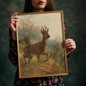 May include: A rectangular framed painting featuring a brown roe deer with antlers standing in a grassy field, with a second smaller deer visible in the background near bare trees. The artwork is enclosed in a thin, metallic gold-coloured frame.