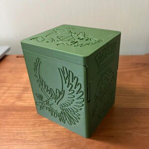 May include: Green, square storage box with embossed designs. The box features an eagle on one side and the word "Pokemon" on another. The top of the box has additional embossed designs. The box is sitting on a wooden surface.