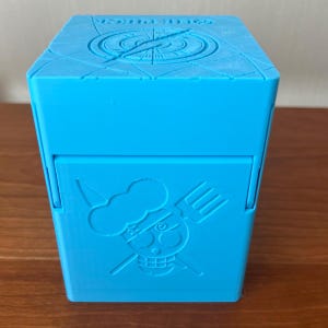 Può includere: Una scatola esagonale blu brillante con coperchio incernierato. Il coperchio presenta un design a bussola. La parte anteriore della scatola mostra un teschio dei cartoni animati con cappello da chef e utensili incrociati. La scatola è in materiale opaco.