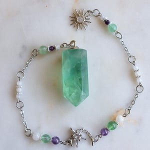 May include: A silver-toned chain necklace featuring a central green hexagonal crystal pendant. The chain is accented with small green, purple, and white beads, a silver sun charm with clear stones, and a silver crescent moon charm with a star.