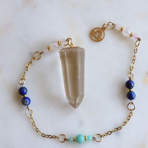 May include: A gold-tone chain bracelet featuring a central faceted smoky quartz crystal pendant, accented with light pink, deep blue, and turquoise beads, and a circular charm displaying the Libra zodiac symbol in a gold-tone finish.