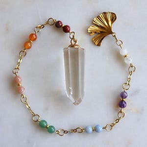 May include: A gold-toned chain bracelet featuring a central clear, faceted crystal point pendant. The chain is accented with pairs of small, multi-colored round gemstone beads and a gold-toned metal ginkgo leaf charm at one end.