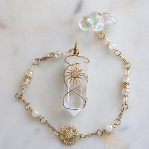 May include: A gold-toned chain bracelet featuring a clear, hexagonal crystal pendant wrapped in gold wire with a sun motif. The chain includes small clear beads, a central sun charm with embedded stones, and an iridescent cloud-shaped charm.