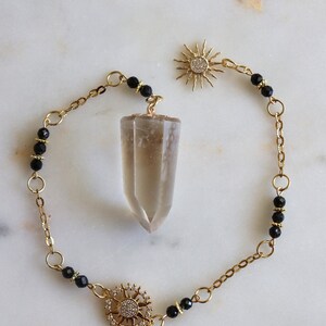 May include: A gold-toned chain necklace featuring a central faceted smoky quartz crystal pendant. The chain is accented with small black beads and two sun-shaped charms, one of which is embellished with tiny clear stones, creating a detailed piece.