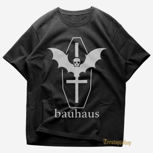Puede incluir: Camiseta negra con un gráfico blanco de un ataúd con alas de murciélago, una calavera y una cruz. La palabra "bauhaus" está impresa debajo en una fuente sans-serif. Cuello redondo.