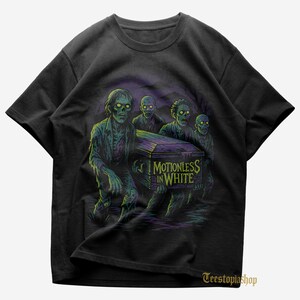 Może przedstawiać: Czarny t-shirt z grafiką czterech zombie niosących trumnę. Na trumnie widnieje napis "Motionless in White". Zombie są zielone i fioletowe, na ciemnym tle.