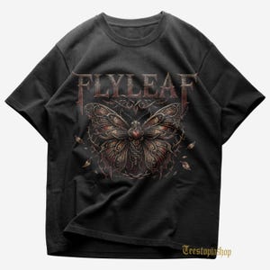Puede incluir: Camiseta negra con el nombre de la banda "FLYLEAF" en fuente gótica sobre un diseño detallado de mariposa. La mariposa tiene patrones intrincados en tonos marrones y rojos, con vides espinosas y hojas. Cuello redondo.