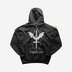Puede incluir: Sudadera con capucha negra con un gráfico blanco en la espalda. El gráfico presenta un diseño de murciélago, calavera, ataúd y cruz, con la palabra "bauhaus" debajo. La sudadera es de color oscuro.