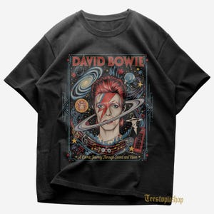 Puede incluir: Camiseta negra con un gráfico colorido de David Bowie, con un rayo en la cara, rodeado de un diseño cósmico. El texto "DAVID BOWIE" está en la parte superior, con "A Cosmic Journey Through Sound and Vision" debajo.