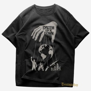 May include: Black t-shirt with a graphic design showing a hand manipulating the Earth with strings. The text "SYSTEM OF A DOWN" is above the globe. Below the globe are illustrations of a person, fire, a bomb, a gun, and a building.