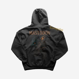 Puede incluir: Sudadera con capucha negra con el nombre de la banda "MASTODON" en letras naranjas. La parte posterior de la sudadera presenta una ilustración detallada de una criatura con acentos naranjas y verde azulado. El nombre de la tienda "Teestupiashop" está impreso en la parte superior derecha.