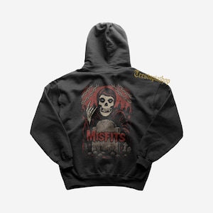 Może przedstawiać: Czarna bluza z kapturem z grafiką postaci z czaszką i napisem "MISFITS" w kolorze czerwonym. Projekt zawiera scenę cmentarza i łańcuchy, z tekstem "Teestopiashop" po prawej stronie u góry.