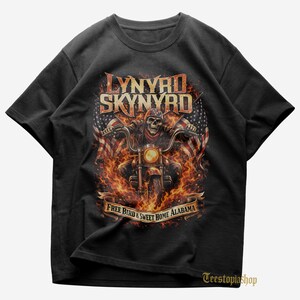 May include: Black t-shirt featuring a graphic of a skull riding a motorcycle engulfed in flames. The design includes the words "LYNYRD SKYNYRD" above and "FREE BIRD & SWEET HOME ALABAMA" below. American flags are on either side of the motorcycle.