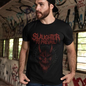 May include: Black t-shirt with the text "SLAUGHTER TO PREVAIL" in red, above a red demonic face graphic. The person in the image is wearing a black cap and blue jeans. The background is a graffiti-covered wall.