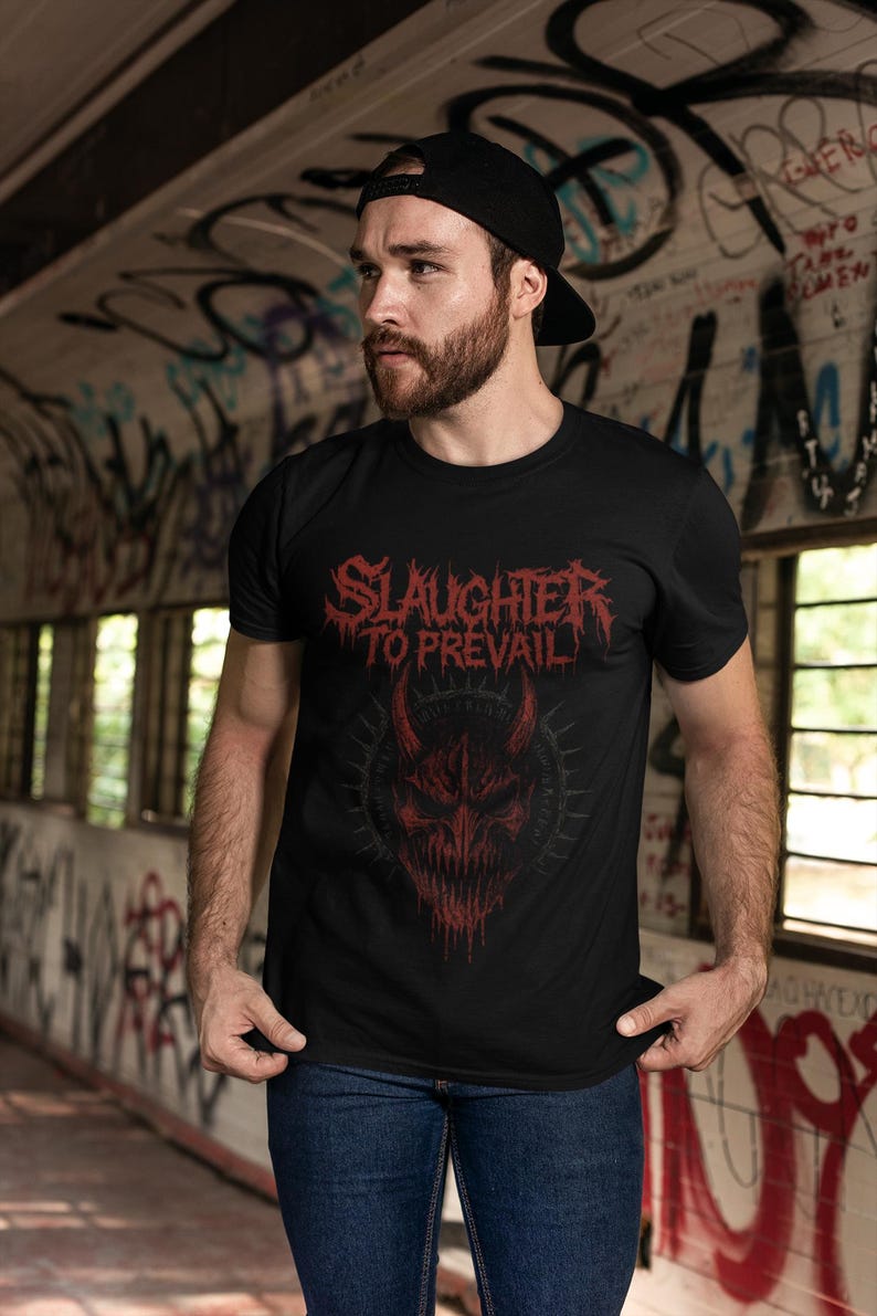 May include: Black t-shirt with the text "SLAUGHTER TO PREVAIL" in red, above a red demonic face graphic. The person in the image is wearing a black cap and blue jeans. The background is a graffiti-covered wall.