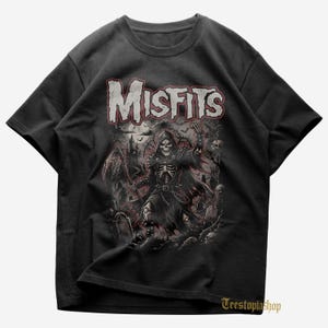 May include: Black t-shirt featuring the word "Misfits" in a distressed, red and white font. The graphic depicts a skeleton in a hooded cloak with a scythe, surrounded by tombstones and a castle. The shirt has a crew neck.
