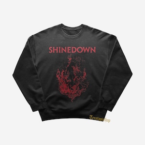 Pode incluir: Uma camisola preta de manga comprida com gola redonda, apresentando a palavra SHINEDOWN em letras maiúsculas vermelhas no topo. Abaixo do texto, há uma ilustração gráfica vermelha de uma figura rodeada por chamas. O texto Teestopiashop está visível.