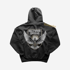 May include: Black hooded sweatshirt featuring a graphic of a dove with outstretched wings, a shield, and the text "HOLLYWOOD UNDEAD". The design is in shades of white, gray, and gold.