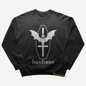 Peut inclure: Sweat-shirt gris foncé avec un motif blanc représentant un cercueil, des ailes de chauve-souris, un crâne et une croix. Le mot "bauhaus" est imprimé en dessous. Le sweat-shirt a un col rond.