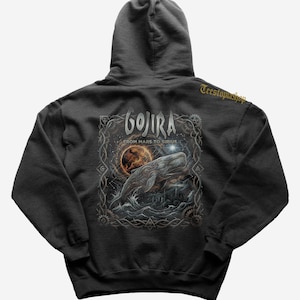 Puede incluir: Sudadera con capucha gris oscuro con un gráfico detallado de una ballena, un planeta y el texto "GOJIRA FROM MARS TO SIRIUS". El diseño está enmarcado con bordes y engranajes ornamentados. La sudadera tiene capucha y bolsillo de canguro.