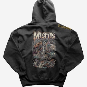 May include: Dark grey hoodie with a graphic of a skeleton playing a guitar, the word "MISFITS" above. The design includes skulls and punk rock imagery. The hoodie has a drawstring hood and is made of a soft material.
