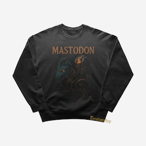 Puede incluir: Sudadera gris oscuro con la palabra "MASTODON" en letras marrones sobre una ilustración detallada de una criatura parecida a un dragón con detalles en naranja. El diseño está centrado en la parte delantera de la sudadera.