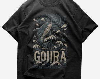 Può includere: T-shirt grigio scuro con una balena che emerge dalle onde e la scritta "GOJIRA". Il design utilizza colori scuri e nuvole vorticose, creando un effetto drammatico.
