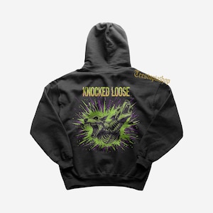 Może przedstawiać: Czarna bluza z kapturem z graficznym wzorem na plecach. Projekt przedstawia warczącego wilka z zielonymi i fioletowymi akcentami oraz złotym napisem "KNOCKED LOOSE".