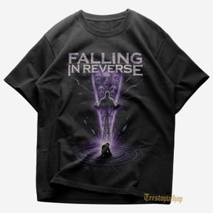 以下が含まれることがあります： 黒いTシャツ。バンド名「Falling in Reverse」が白でプリントされています。グラフィックデザインは、紫と白の色合いで、人、塔、顔が描かれています。ダークでゴシックなテーマです。