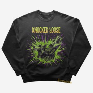 Może przedstawiać: Ciemnoszara bluza z nadrukiem graficznym przedstawiającym dwa warczące stworzenia podobne do wilków z zielonymi i fioletowymi akcentami. Tekst "KNOCKED LOOSE" jest wyświetlany na żółto.