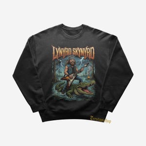 May include: A black crewneck sweatshirt featuring a graphic of a bearded guitarist wearing a bandana and vest, playing a V-shaped electric guitar while riding a large green alligator in a swampy setting with lightning, under the text Lynyrd Skynyrd.