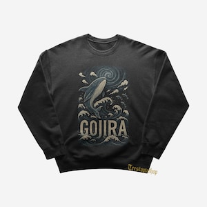 May include: Dark gray sweatshirt featuring a blue whale breaching through ocean waves, with the word "GOJIRA" in a vintage font. The design includes swirling clouds and a stylized ocean scene. The sweatshirt has a crew neck.