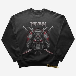 May include: Dark gray sweatshirt featuring a graphic of a samurai skull with crossed swords and the band name "Trivium" in silver. The design includes red accents and intricate details, creating a bold and edgy aesthetic.
