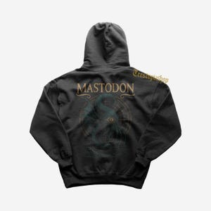 Puede incluir: Sudadera con capucha gris oscuro con la palabra "MASTODON" en letras doradas sobre un gráfico verde azulado y dorado de una criatura marina. La sudadera con capucha tiene una capucha y mangas largas. El nombre de la tienda Etsy "Teestupiashop" está impreso en oro en la parte superior derecha.