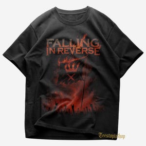 May include: Black t-shirt featuring the band name "FALLING IN REVERSE" in red letters above a flaming flag design. The flag has a crown and an "X" symbol. A crowd with raised hands is visible below the flag.