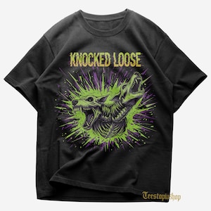 Może przedstawiać: Czarny t-shirt z grafiką przedstawiającą dwa warczące stworzenia podobne do wilków z odsłoniętymi klatkami piersiowymi, na tle zielono-fioletowego wzoru. Tekst "KNOCKED LOOSE" jest wydrukowany nad grafiką w postarzonej, złotej czcionce.