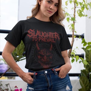May include: Black t-shirt with the text "SLAUGHTER TO PREVAIL" in red, above a red demonic face graphic. The model is wearing a denim skirt and is standing outside.
